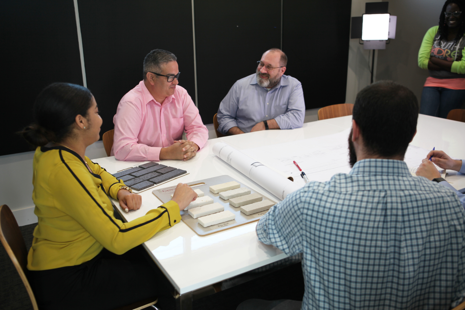 A group of four people is gathered around a table in a modern office setting. The person on the left, wearing a yellow shirt with black stripes, is pointing toward a selection of sample materials laid out on a board. There are several rectangular samples in various shades of gray and white. The man in a light pink shirt is seated next to her, looking attentively at the materials. To the right, another man with a beard is smiling while engaging in the discussion, and a third man in a blue checkered shirt is writing notes on a roll of plans that is partially visible on the table. In the background, a woman with long hair, wearing a green shirt, stands near a light source, observing the group. The walls are primarily black, providing a contrasting backdrop to the discussion.